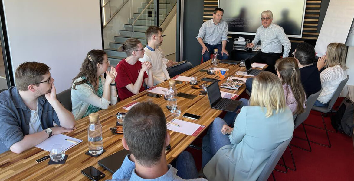 L'image montre une réunion en cours dans une salle de conférence moderne. Plusieurs personnes sont assises autour d'une grande table en bois, équipées de laptops, de bouteilles d'eau et de documents. Deux hommes sont debout et semblent animer la réunion ou faire une présentation. L'un d'eux est près d'un écran ou tableau, ce qui suggère qu'il pourrait être en train de partager des informations ou des données avec le groupe. Les participants semblent attentifs et engagés dans la discussion. La salle est bien éclairée, avec de grandes fenêtres laissant entrer la lumière naturelle, et elle est décorée de manière professionnelle.